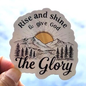 May include: A sticker with a mountain and sunrise design. The text reads "Rise and shine & give God the Glory" in black script. The sticker is held by a hand.