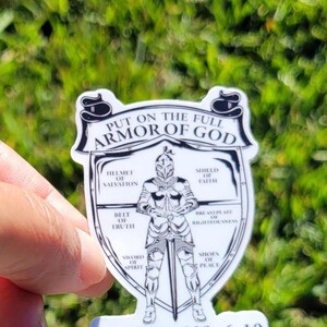 May include: A white sticker with a black and white illustration of a knight wearing armor. The knight is standing in the center of a shield. The shield is surrounded by a circle with the words "Put on the full armor of God" at the top and "Ephesians 6:10" at the bottom. The circle is divided into eight sections, each with a different part of the armor of God listed.
