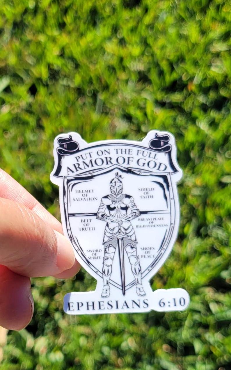 May include: A white sticker with a black and white illustration of a knight wearing armor. The knight is standing in the center of a shield. The shield is surrounded by a circle with the words "Put on the full armor of God" at the top and "Ephesians 6:10" at the bottom. The circle is divided into eight sections, each with a different part of the armor of God listed.