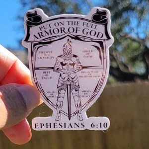 May include: A white sticker with a black outline of a knight in armor. The knight is holding a sword and is surrounded by a shield with the words "Put on the full armor of God" and "Ephesians 6:10". The shield is divided into sections with the names of the different pieces of armor: helmet of salvation, shield of faith, breastplate of righteousness, belt of truth, sword of the spirit, and shoes of peace.