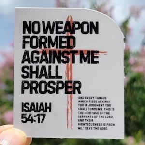May include: A white sticker with the text "NO WEAPON FORMED AGAINST ME SHALL PROSPER" and "ISAIAH 54:17" in black. A red cross is in the center. Additional text is at the bottom. The sticker is held up against a blurred background.