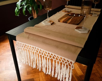 Op de afbeelding: Een beige tafelloper met witte franjes, op een zwarte tafel. De loper wordt gecombineerd met heldere wijnglazen, onderzetters en een houten snijplank. De tafel is gedekt voor een diner.