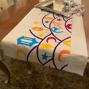 Op de afbeelding: Een witte tafelrunner met geborduurde halve manen, sterren en lantaarns in tinten blauw, geel, oranje, rood en paars. De runner ligt op een houten tafel met een zilveren dienblad.