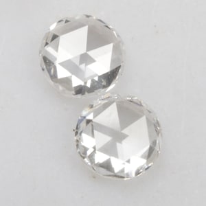 Puede incluir: Dos gemas redondas y transparentes con superficies facetadas. Las gemas son incoloras y reflejan la luz, mostrando sus intrincados cortes. Las piedras están sobre un fondo blanco, resaltando su brillo. Podrían usarse para joyería.
