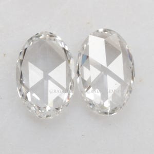 Puede incluir: Dos piedras preciosas transparentes de forma ovalada con facetas de corte rosa. Cada piedra presenta una base plana y una parte superior abovedada con facetas triangulares que reflejan la luz. El texto @RADHAGEMSINDIA es visible.