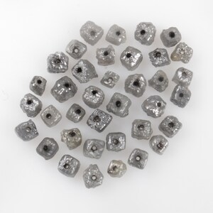 May include: A collection of rough-cut, grey diamond beads, each with a drilled hole. The beads have an irregular shape and a metallic sheen. The beads are scattered on a white surface.
