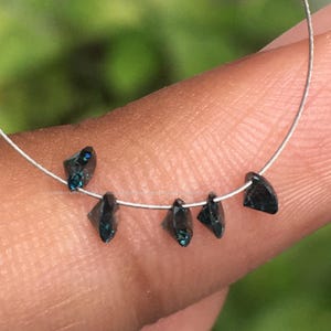 May include: A strand of dark blue faceted gemstone beads strung on a silver wire. The beads are triangular in shape and have a slight green hue.