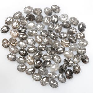 Pode incluir: Um monte de diamantes cinzentos soltos, de forma oval, com inclus&otilde;es pretas. Os diamantes est&atilde;o espalhados sobre uma superf&iacute;cie branca.