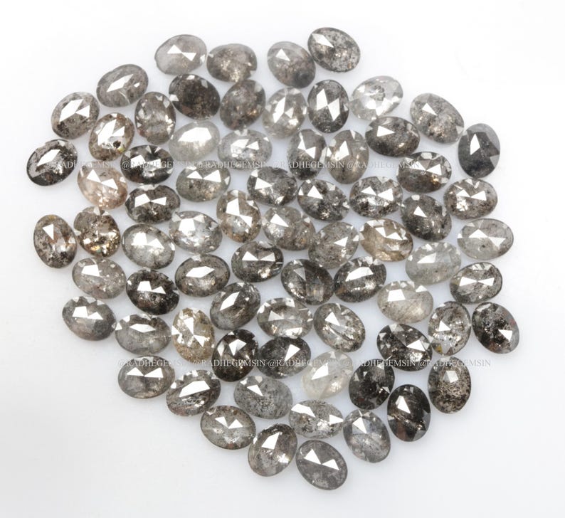 Pode incluir: Um monte de diamantes cinzentos soltos, de forma oval, com inclus&otilde;es pretas. Os diamantes est&atilde;o espalhados sobre uma superf&iacute;cie branca.