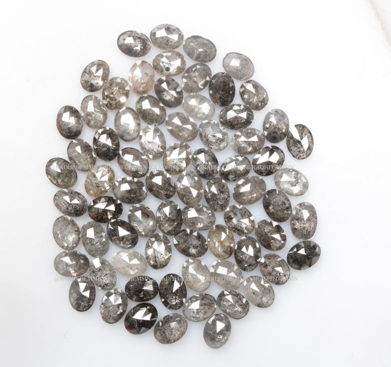 Pode incluir: Uma cole&ccedil;&atilde;o de diamantes cinzentos soltos, de forma oval, com um acabamento &aacute;spero e n&atilde;o cortado. Os diamantes est&atilde;o espalhados sobre uma superf&iacute;cie branca.