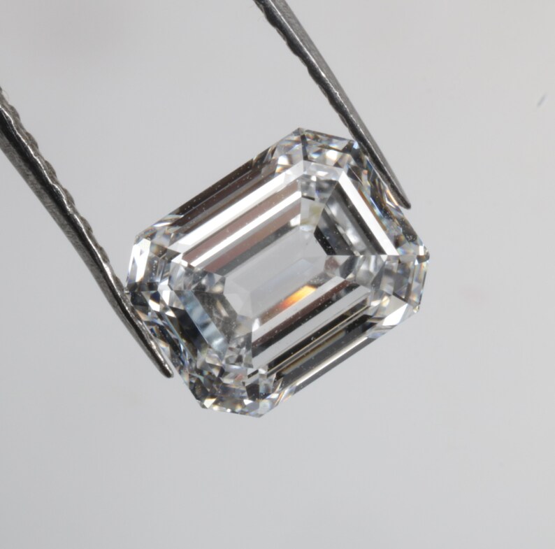 Pu&ograve; includere: Una gemma di diamante taglio smeraldo trasparente tenuta da una pinzetta.