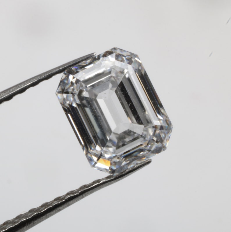 Pu&ograve; includere: Una gemma di diamante trasparente a taglio smeraldo tenuta da una pinzetta.