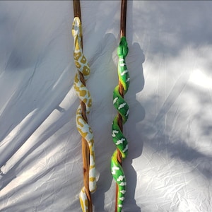 以下が含まれることがあります： 2本の木製の杖で、それぞれ詳細な手作りのヘビのデザインで飾られています。1匹のヘビは黄色と白、もう1匹は緑と白です。ヘビは木の杖に巻き付いています。