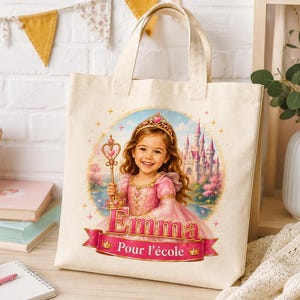 Peut inclure: Un sac fourre-tout en toile couleur crème avec une illustration imprimée d'une jeune enfant portant une robe rose et un diadème doré, tenant une baguette à embout en forme de cœur. Le texte indique Emma et Pour l'école sur une bannière rose.