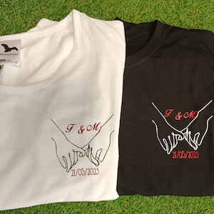 Peut inclure: Deux t-shirts blancs et noirs avec un motif brodé rouge de deux mains se tenant l'une l'autre avec le texte "F & M 21/05/2023" sur chaque t-shirt.