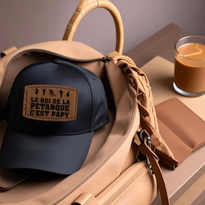 Peut inclure: Casquette de baseball noire avec un patch en cuir marron portant l'inscription "LE ROI DE LA PETANQUE C'EST PAPY" et des silhouettes de joueurs de pétanque. La casquette est dans un sac beige, avec un café et un portefeuille marron.
