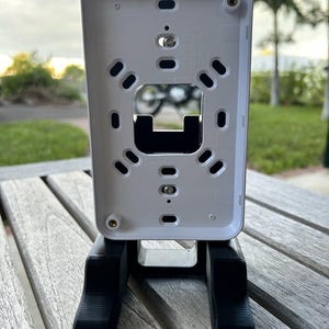 Könnte beinhalten: Ein weißes rechteckiges Gerät mit einer Reihe von Löchern und einem zentralen Ausschnitt, montiert auf einem schwarzen, rechteckigen Ständer. Das Gerät steht auf einem Holztisch mit verschwommenem Hintergrund im Freien.