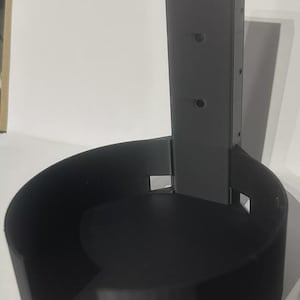 Puede incluir: Un soporte de pared negro, impreso en 3D, para un dispositivo doméstico inteligente. El soporte tiene una base circular y un soporte vertical con tres agujeros para el montaje. El diseño es simple y funcional, adecuado para organizar y mostrar tecnología.