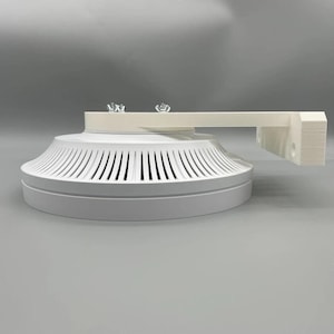 Può includere: Un dispositivo bianco e circolare per montaggio a soffitto con una serie di fessure verticali sul perimetro. Ha un braccio rettangolare che si estende verso destra, con due viti sulla parte superiore. Il dispositivo è progettato per il montaggio.