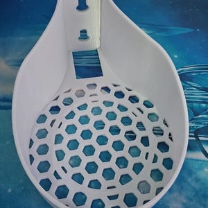 May include: A white, 3D-printed wall-mounted basket with a honeycomb-patterned base. The basket has a teardrop shape and a bracket with two keyhole slots for hanging. The interior is visible through the hexagonal cutouts.