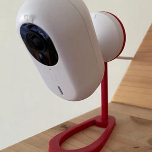 Puede incluir: Una cámara de seguridad blanca con una lente negra montada en un soporte rojo. El soporte tiene una base curva y un soporte vertical. La cámara está colocada sobre una superficie de madera.