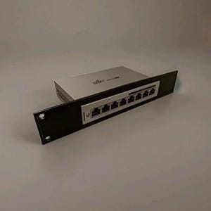 Könnte beinhalten: Ein schwarzes Rack-Mount mit acht Ethernet-Anschlüssen und ein silbernes Netzwerkgerät. Das Gerät trägt den Markennamen "Ubiquiti". Das Rack-Mount hat zwei Befestigungslöcher.