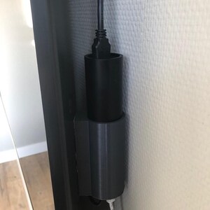 Op de afbeelding: Een zwart, cilindrisch apparaat met een USB-kabel erin, gemonteerd op een witte muur. Het apparaat lijkt een houder of adapter te zijn, mogelijk 3D-geprint, ontworpen om de kabel te organiseren of vast te zetten.