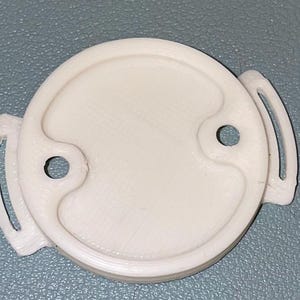 Könnte beinhalten: Ein weißes, kreisförmiges Objekt mit zwei kleinen Löchern und zwei gebogenen Griffen. Das Objekt hat eine strukturierte Oberfläche und scheint aus Kunststoff zu bestehen. Das Design ist symmetrisch, mit einem erhöhten Mittelbereich.