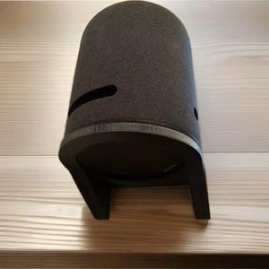 May include: A dark gray fabric-covered speaker with a black base and stand. The speaker has a curved, cylindrical shape with a horizontal slot and a small hole. The stand is a U-shaped design, providing stability.