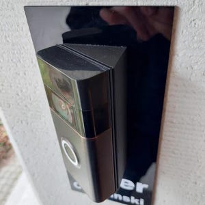 Puede incluir: Un timbre de video negro montado en una placa rectangular negra. El timbre tiene un borde de color bronce y un botón plateado. La placa está montada en una pared texturizada gris claro. El texto "er nski" es visible.