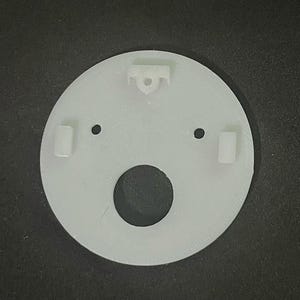 May include: A white, circular plastic component with a large central hole and two smaller holes. It features two rectangular protrusions and a hook-shaped element. The object is designed for a specific mechanical or structural purpose.