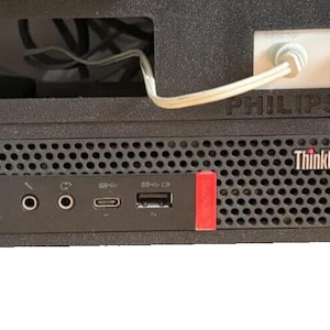 Può includere: Un case per computer nero con un computer ThinkCentre. Il pannello frontale presenta varie porte e una copertura rossa per la porta USB. Il testo "PHILIPS HUE" è visibile sopra il computer.
