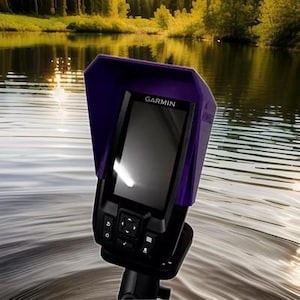 May include: A black Garmin fish finder with a purple visor is mounted in the water. The device has a screen and several buttons. The background shows a lake with trees and sunlight reflecting on the water.