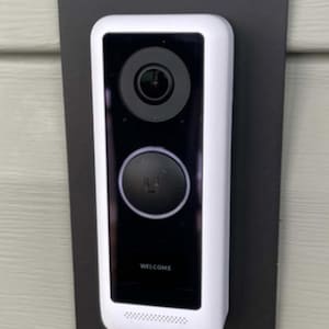 Puede incluir: Un timbre inteligente blanco y negro con una lente de cámara y un botón. El timbre está montado en una placa rectangular negra y está fijado a una pared verde claro. La palabra "WELCOME" está impresa en el timbre.