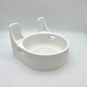 May include: A white, 3D-printed wall mount for a smart home device. The mount features a circular recess and two raised sections with mounting holes. The design is simple and functional, with a clean, modern aesthetic.