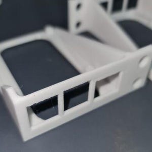 Puede incluir: Varios componentes de plástico blanco impresos en 3D están dispuestos sobre una superficie gris. Cada componente tiene un marco rectangular con tres aberturas y una superficie oscura, posiblemente reflectante. El diseño incluye soportes angulados y agujeros circulares.
