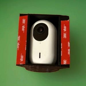 Puede incluir: Una cámara de seguridad blanca, de forma ovalada, con una lente negra y una pequeña luz indicadora. La cámara está montada en un soporte negro con tiras adhesivas rojas 3M VHB. El fondo es verde liso.