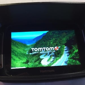 May include: A black GPS device with a screen displaying a scenic road view, framed by a black visor. The screen shows the TomTom logo and the text "Great Rides Edition".
