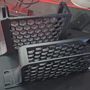 May include: Two black 3D-printed components with a honeycomb pattern. Each piece has a rectangular frame with a series of hexagonal openings. The components have mounting tabs with holes for screws or bolts. The parts are likely for a computer case or other electronic device.