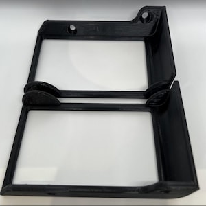 May include: Two black, right-angled brackets, possibly for shelving or mounting. Each bracket features a rectangular frame with rounded corners and two small screw holes. The brackets are stacked, revealing their identical design and construction.