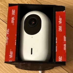 May include: A white security camera with a black lens and a small black microphone hole is mounted inside a black rectangular frame. Red adhesive strips are visible on the frame. A white cable extends from the bottom.