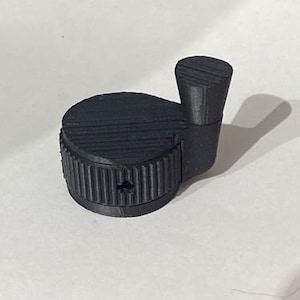 May include: A black, 3D-printed knob with a cylindrical base and a smaller, rectangular handle. The base has a ridged texture, and the handle is also textured. The object is likely a component for a machine or device.