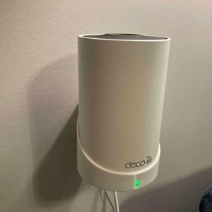 May include: A white cylindrical Deco 6E wireless mesh system mounted on a wall. The device has a curved base and a black top. A green light is illuminated at the bottom, and a power cord is visible.