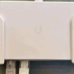 May include: A white electronic device with a rectangular shape and rounded corners, featuring a subtle 'U' logo. The device is mounted on a wall, with cables extending from the bottom. The device is likely a network device.