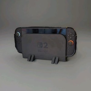 Puede incluir: Una consola Nintendo Switch negra colocada en una base de carga negra. La base tiene el logotipo de Nintendo Switch y dos patas de soporte. La consola tiene dos mandos desmontables con detalles en azul y naranja.