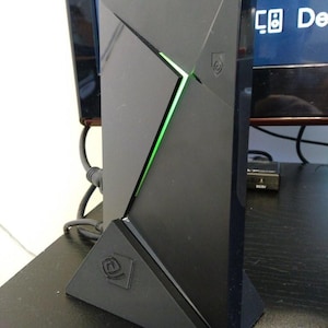 May include: A black, angular media streaming device with a green illuminated accent. The device is standing upright on a triangular base, featuring a logo. The design is modern and geometric, with a sleek, minimalist aesthetic.