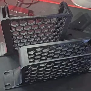 以下が含まれることがあります： 黒色の3Dプリントされたコンピューターケース部品が2つ。各部品はハニカム模様と取り付けタブが付いた長方形のフレームを持っています。コンピューターケース内に収まるように設計されています。