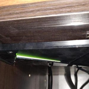 Op de afbeelding: Een zwart elektronisch onderdeel, mogelijk een harde schijf, gemonteerd onder een houten oppervlak. Het apparaat wordt op zijn plaats gehouden door metalen beugels. Een groen licht is zichtbaar en er zijn draden aangesloten.