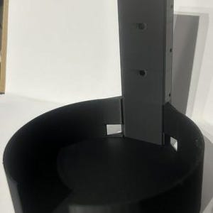 Puede incluir: Soporte de pared negro impreso en 3D para un dispositivo doméstico inteligente. El soporte tiene una base circular con un soporte rectangular vertical con orificios de montaje. El diseño es simple y funcional.