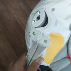 Op de afbeelding: Close-up van een baby wipstoel met een wit en grijs patroon. De wipstoel heeft een lichtgrijs plastic frame en is gedeeltelijk gemonteerd. De stof heeft gele en oranje accenten.